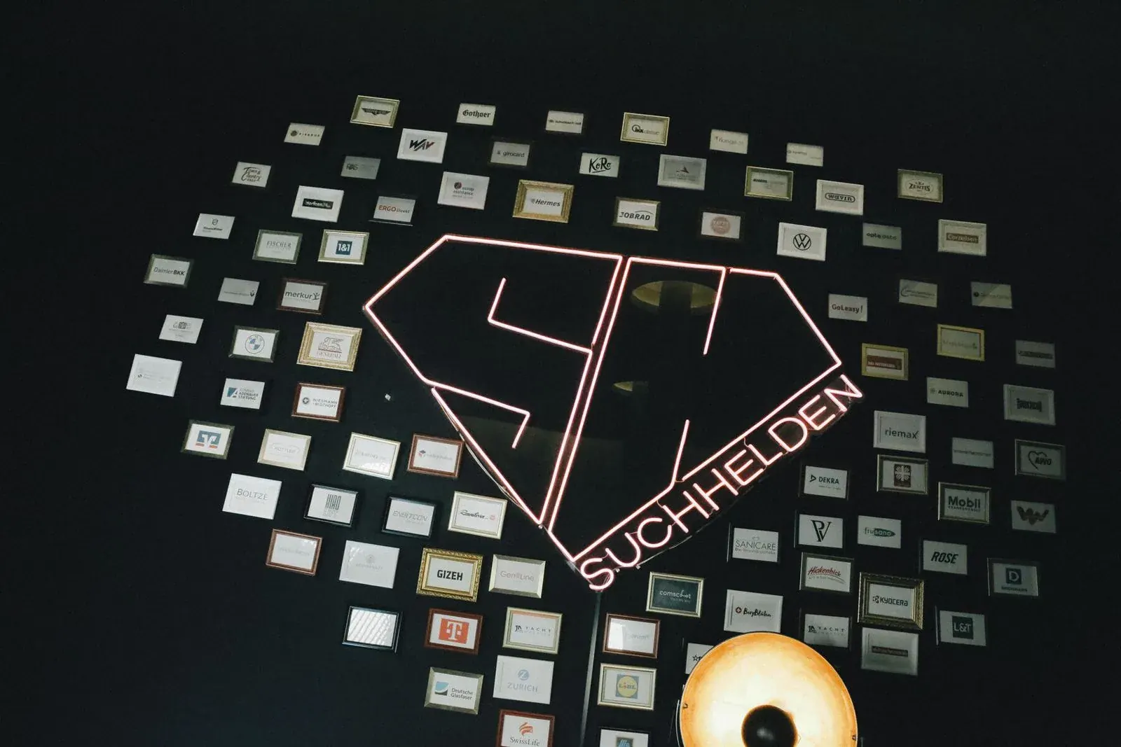 Kundenwand mit gerahmten Logos rund um das SH-Neonlogo im Suchhelden-BĂŒro in OsnabrĂŒck Kundenwand mit gerahmten Logos rund um das SH-Neonlogo im Suchhelden-BĂŒro in OsnabrĂŒck