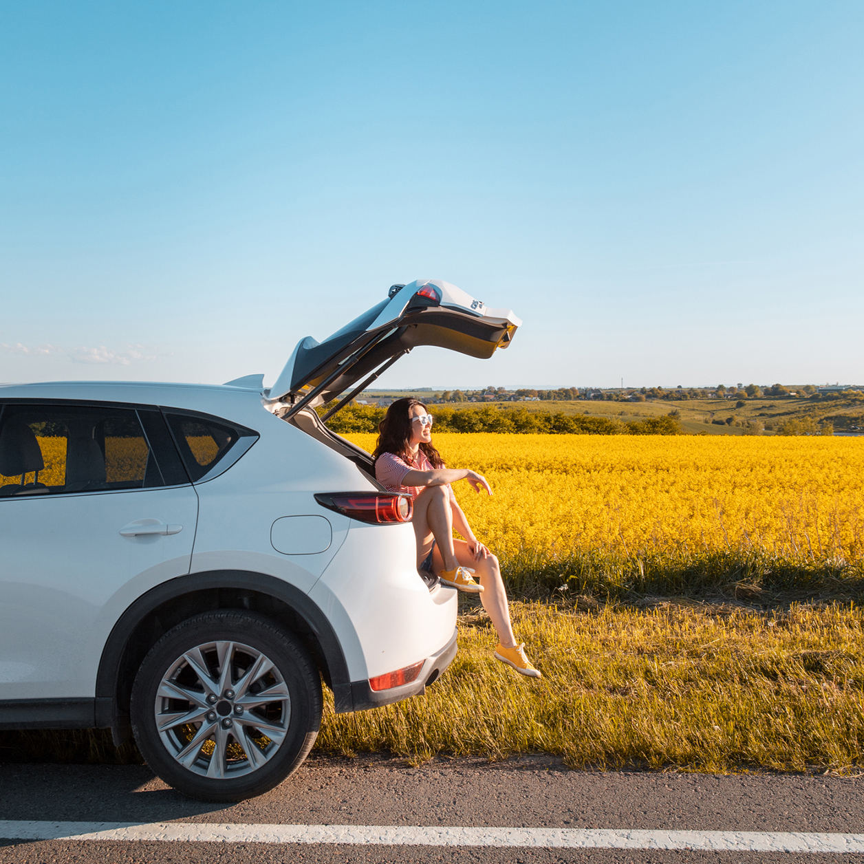 Lächelnde huebsche Frau, die bei Sonnenuntergang im Kofferraum des SUV sitzt und auf das Farmfeld schaut