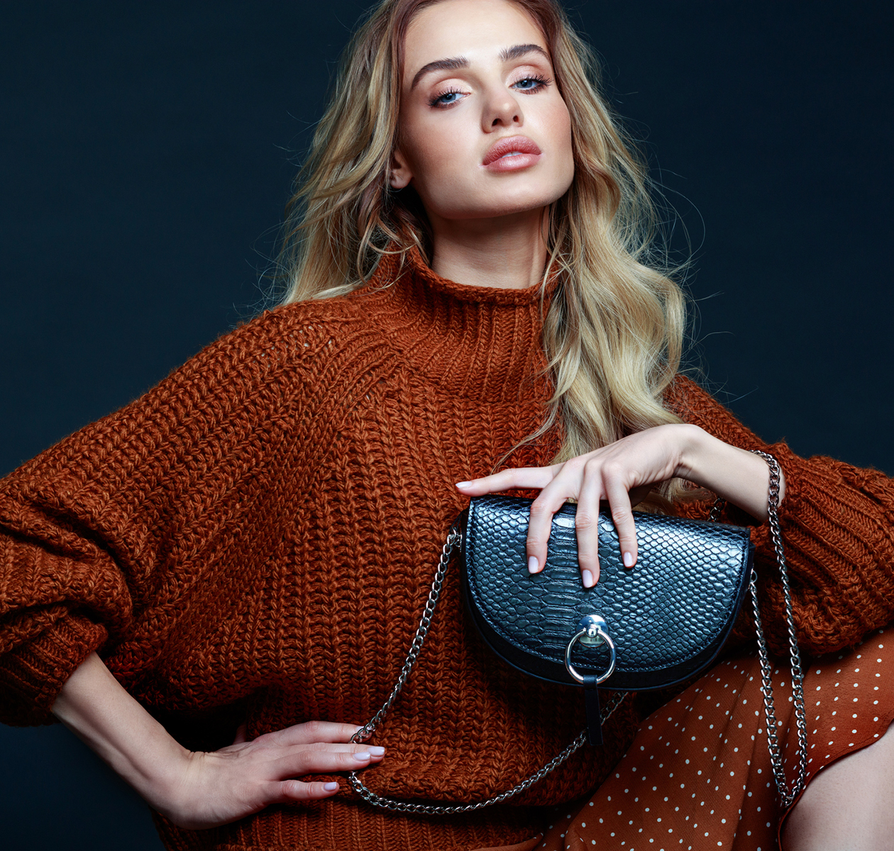 Portraet einer blonden jungen Frau mit langen Haaren, die braunen Pullover und Rock traegt, eine schwarze Handtasche haelt und in die Kamera schaut